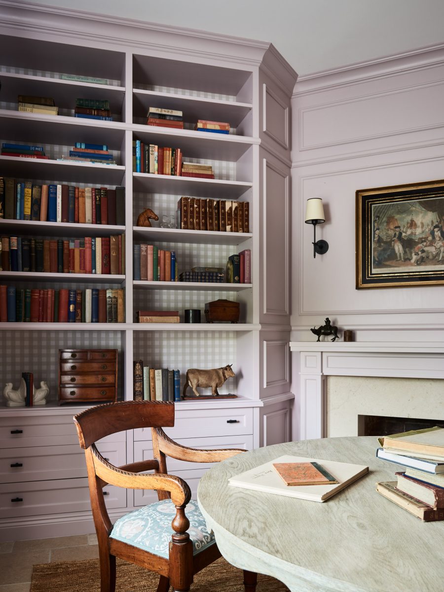 Floor-To-Ceiling Dusty Rose Bookshelves With A Checked Wallpaper Backing, An Antique Timber Chair, And A Round Table With Vintage Books.