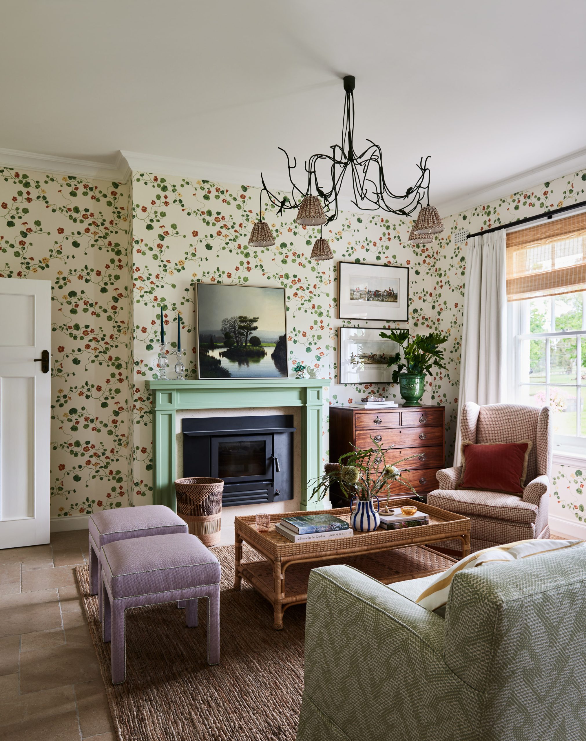 A Living Room Featuring Lake August Nasturtium Wallpaper, A Pale Green Mantelpiece With A Fireplace Insert, A Wicker Coffee Table, And A Black Bird-Themed Chandelier.