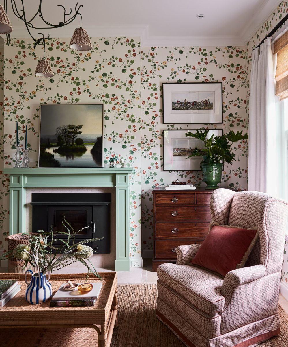 A Pale Green Mantelpiece With A Black Fireplace Insert, Surrounded By Floral Lake August Wallpaper, An Antique Timber Chest, And A Pink Wingback Chair.