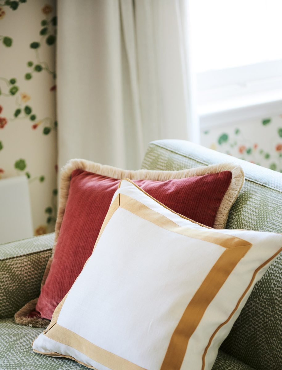 Close-Up Of A Dusty Red Corduroy Cushion With Fringe And A White Cushion With Yellow Trim On A Green Textured Sofa Against Floral Wallpaper.