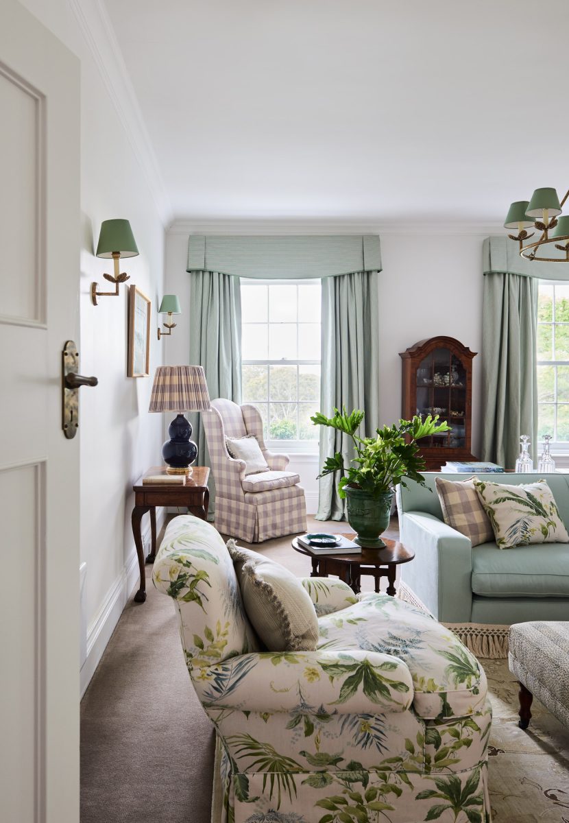A Bright Formal Sitting Room With Large Windows, Light Blue Curtains With Pelmets, A Floral Upholstered Armchair, And An Antique Timber Display Cabinet.