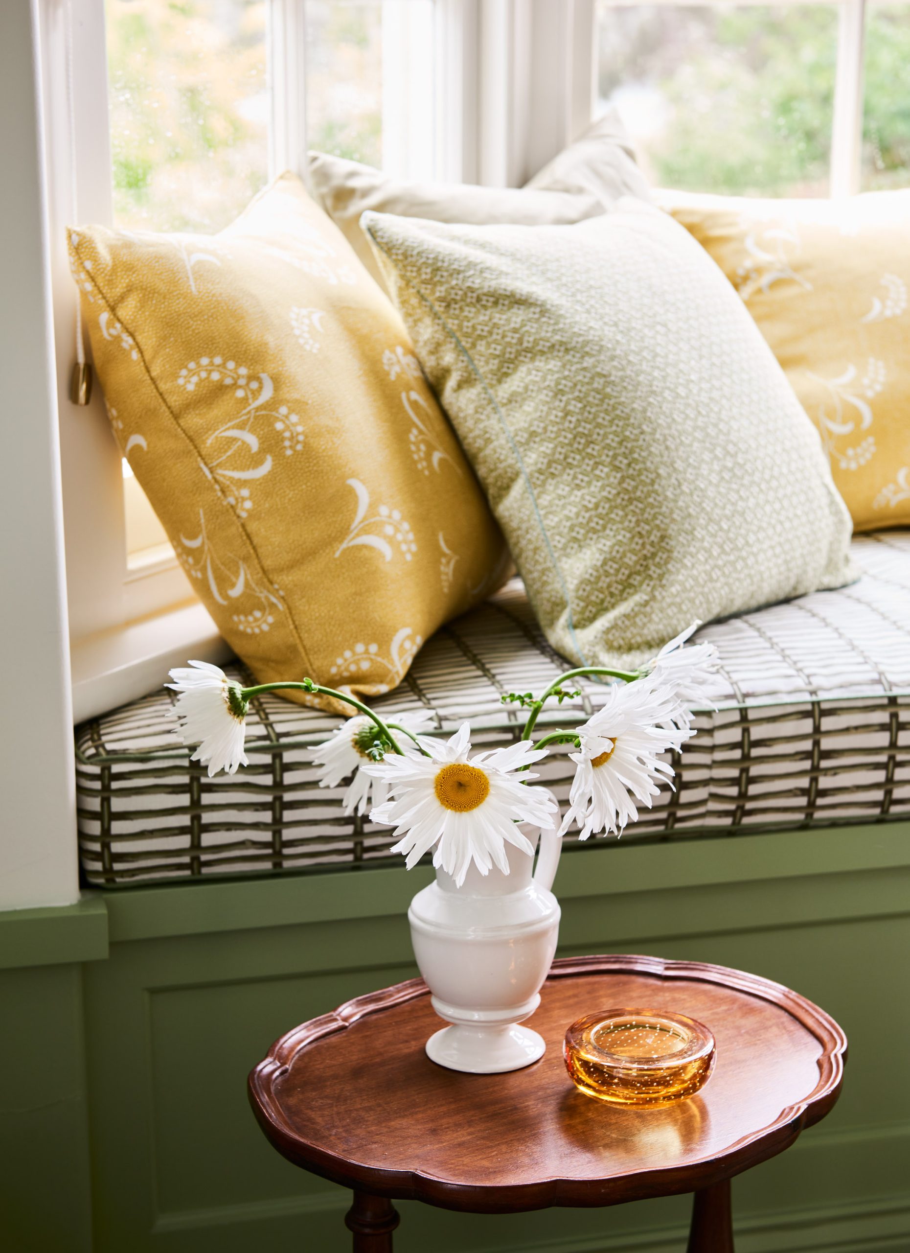 Close-Up Of A Sage Green Painted Window Seat With Yellow Floral And Green Textured Cushions, Beside A Timber Table With A White Vase Of Daisies.