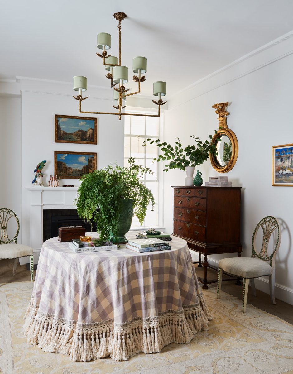 A Round Table With A Purple And White Checkered Fringed Cloth, A Large Green Fern, And An Antique Timber Chest Beneath A Bronze Colombier Chandelier.