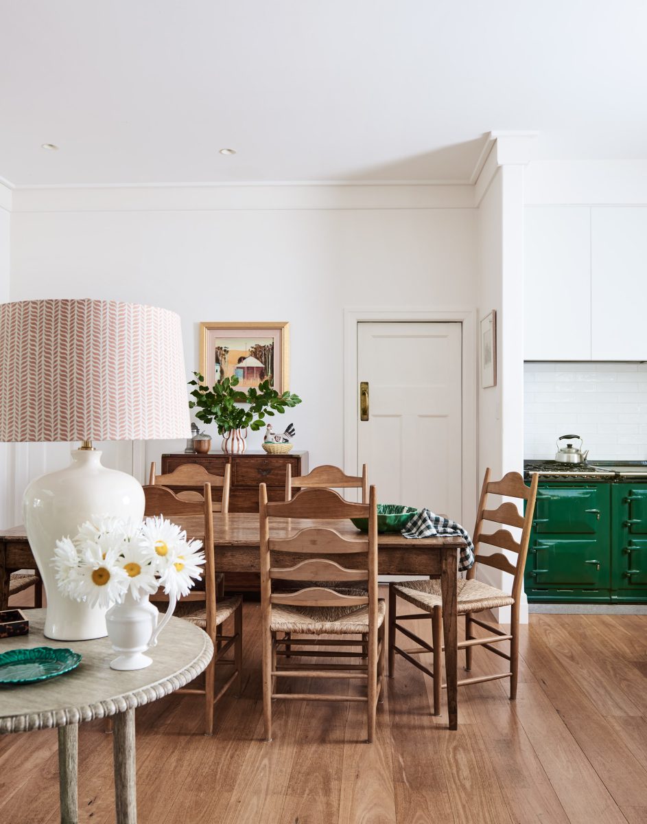 A Warm Heritage Kitchenette Featuring A Vibrant Forest Green Aga Stove, Timber Dining Table With Ladder-Back Chairs, And A White Tiled Splashback.
