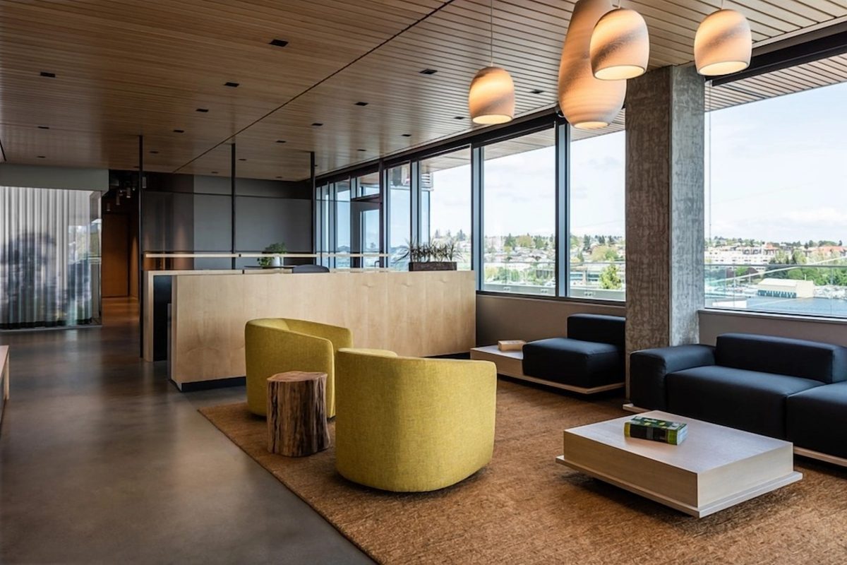 A Modern Commercial Office Breakout Area Featuring A Linear Timber Slat Ceiling, Exposed Concrete Columns, And Expansive Glazing Overlooking A Coastal Urban Landscape.