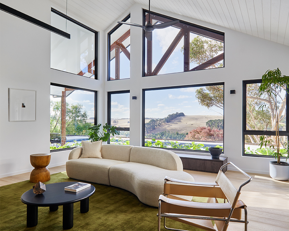 Expansive Architectural Living Area Featuring Vaulted Ceilings, Exposed Timber Trusses, And Large Black-Framed Glazing Overlooking Rural Hills.