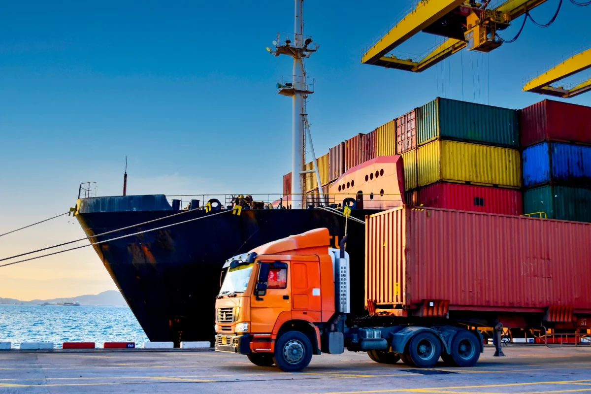 A Large Container Ship At A Port With An Orange Truck In The Foreground, Representing The Complex Global Supply Chain And Freight Challenges Facing The Australian Construction Industry In 2026.