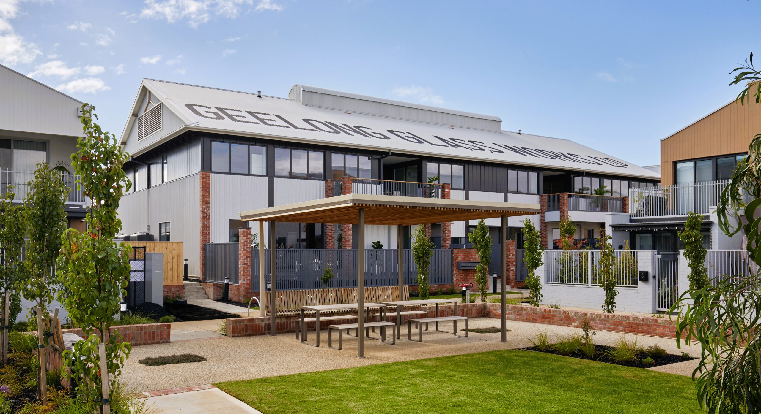 Converted Heritage Warehouse Apartments By Fox Building Group In South Geelong, Showing A Communal Lawn, Timber Pavilion, And Historic Roof Sign.